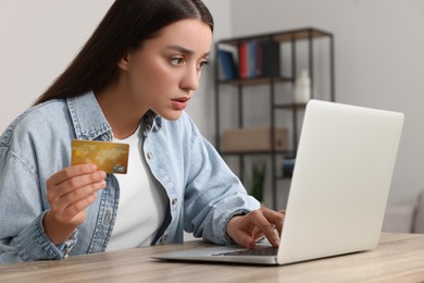 Photo of Confused woman with credit card using laptop at table indoors. Be careful - fraud