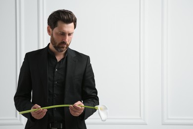 Photo of Sad man with calla lily flower near white wall, space for text. Funeral ceremony