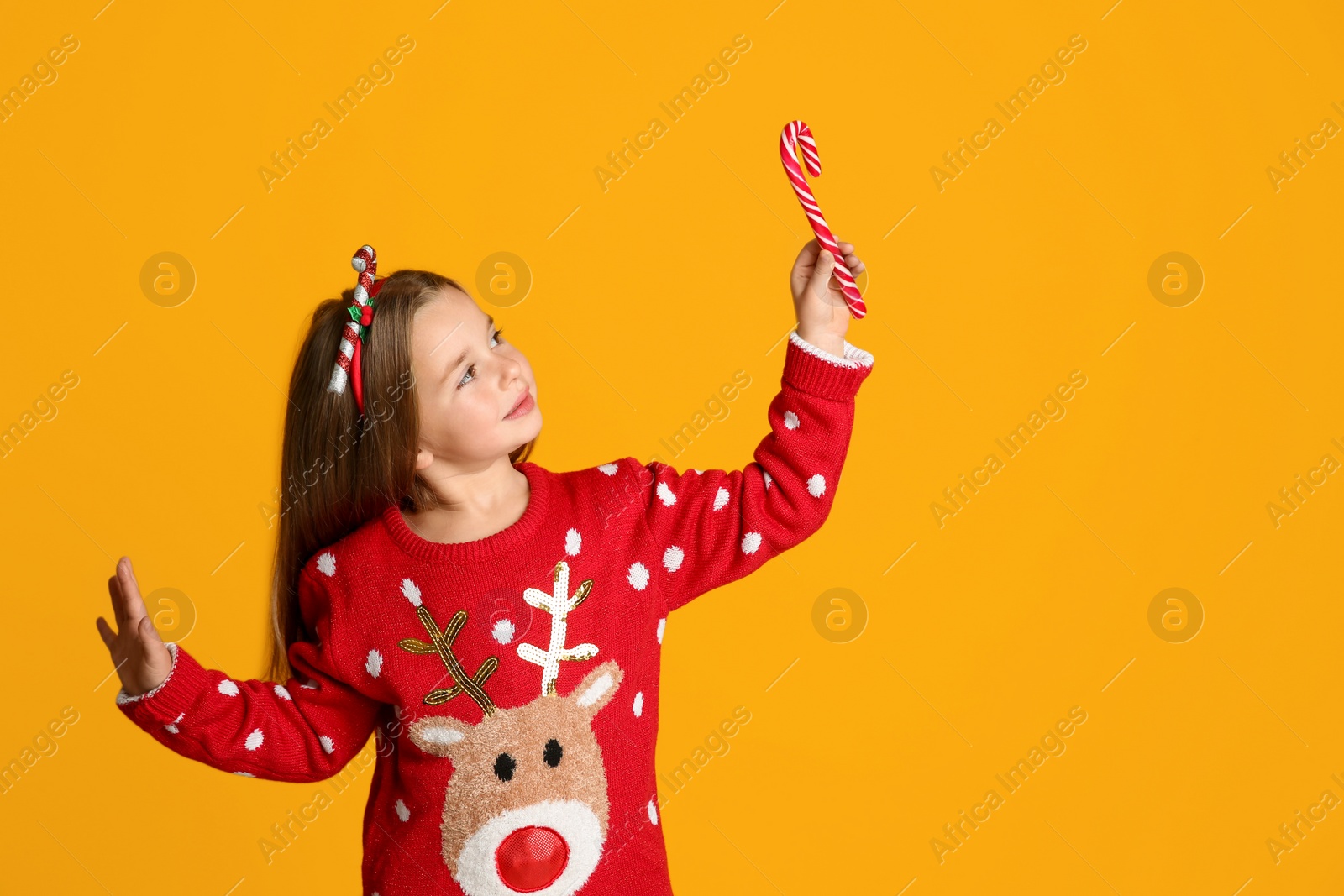 Photo of Cute little girl in Christmas sweater and festive headband holding candy cane on yellow background, space for text