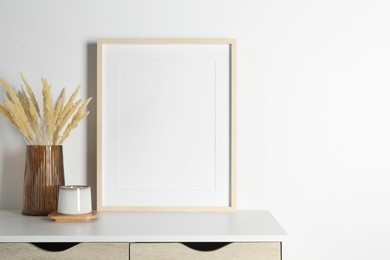 Photo of Empty photo frame, cup and vase with dry decorative spikes on white table. Mockup for design