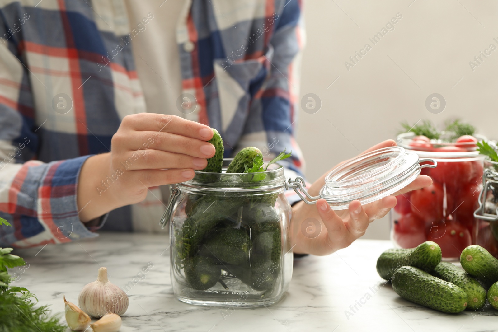 Photo of Woman putting cucumber into glass jar at white marble kitchen table, closeup. Pickling vegetables