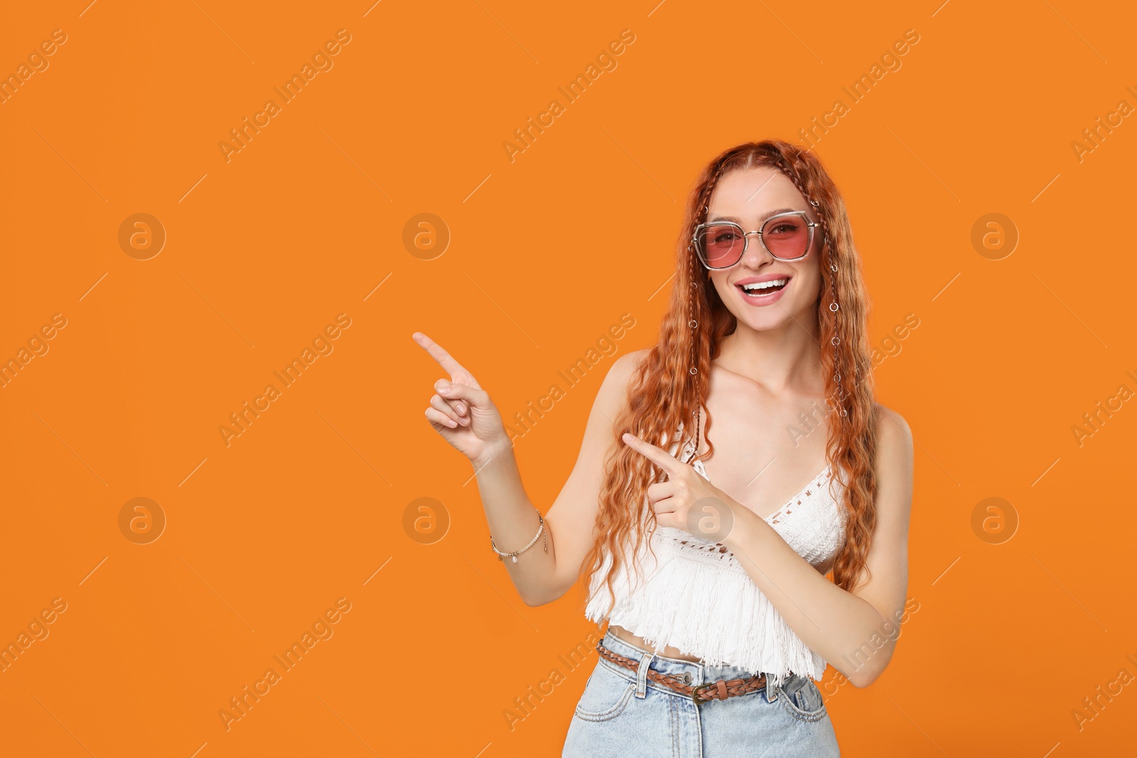 Photo of Stylish young hippie woman in sunglasses pointing at something on orange background, space for text