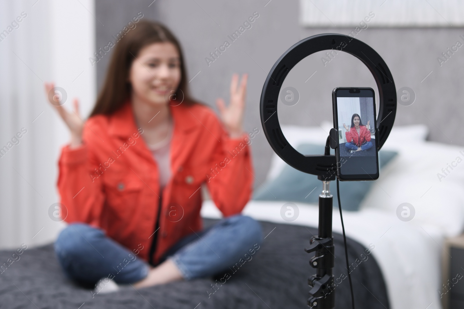 Photo of Smiling teenage blogger explaining something while streaming at home, focus on smartphone