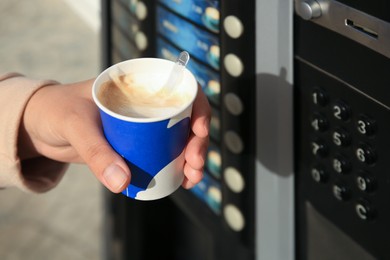 Woman with paper cup of cappuccino near coffee vending machine, closeup