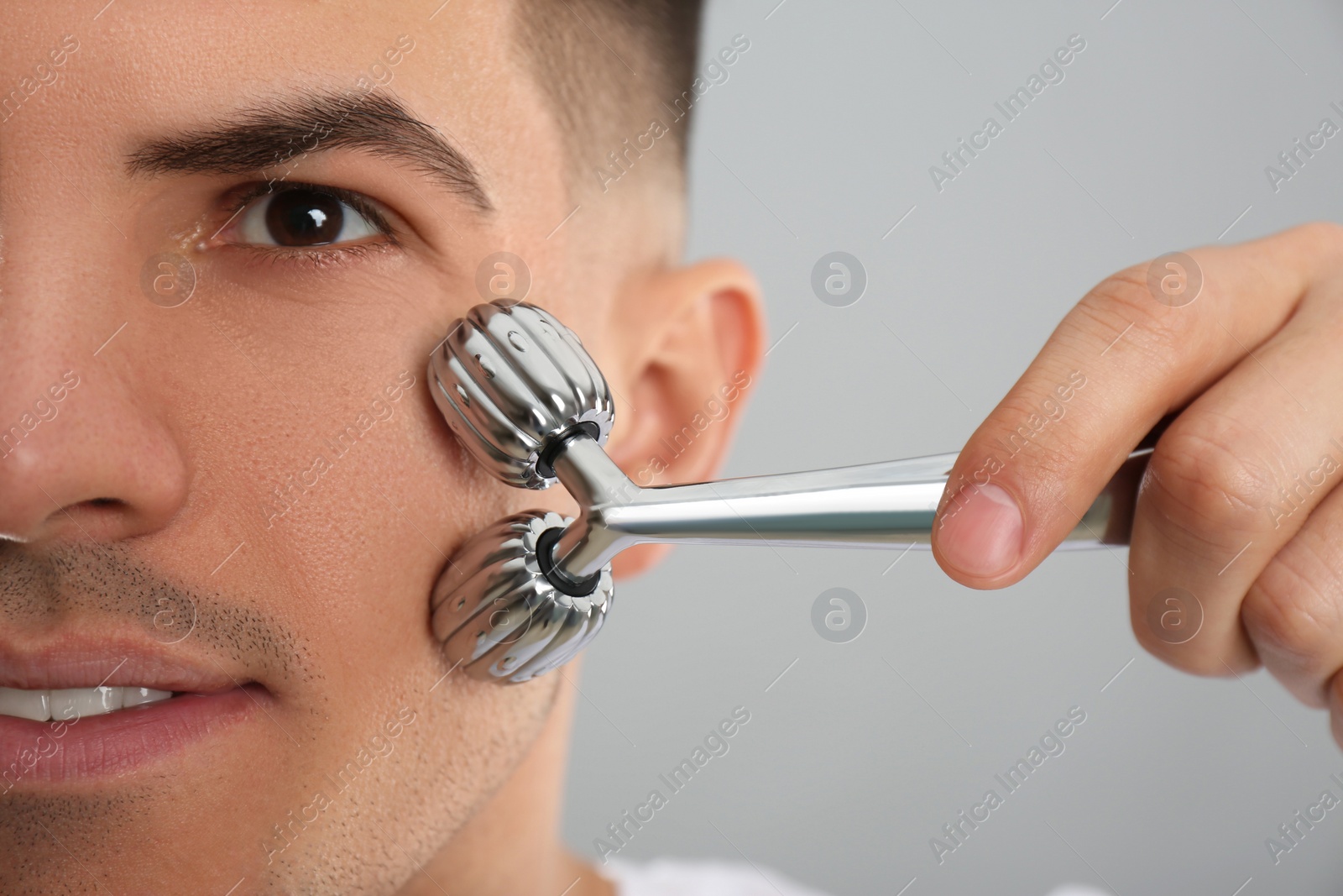 Photo of Man using metal facial roller on grey background, closeup