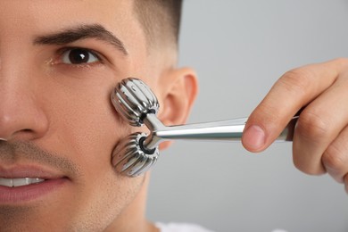 Man using metal facial roller on grey background, closeup