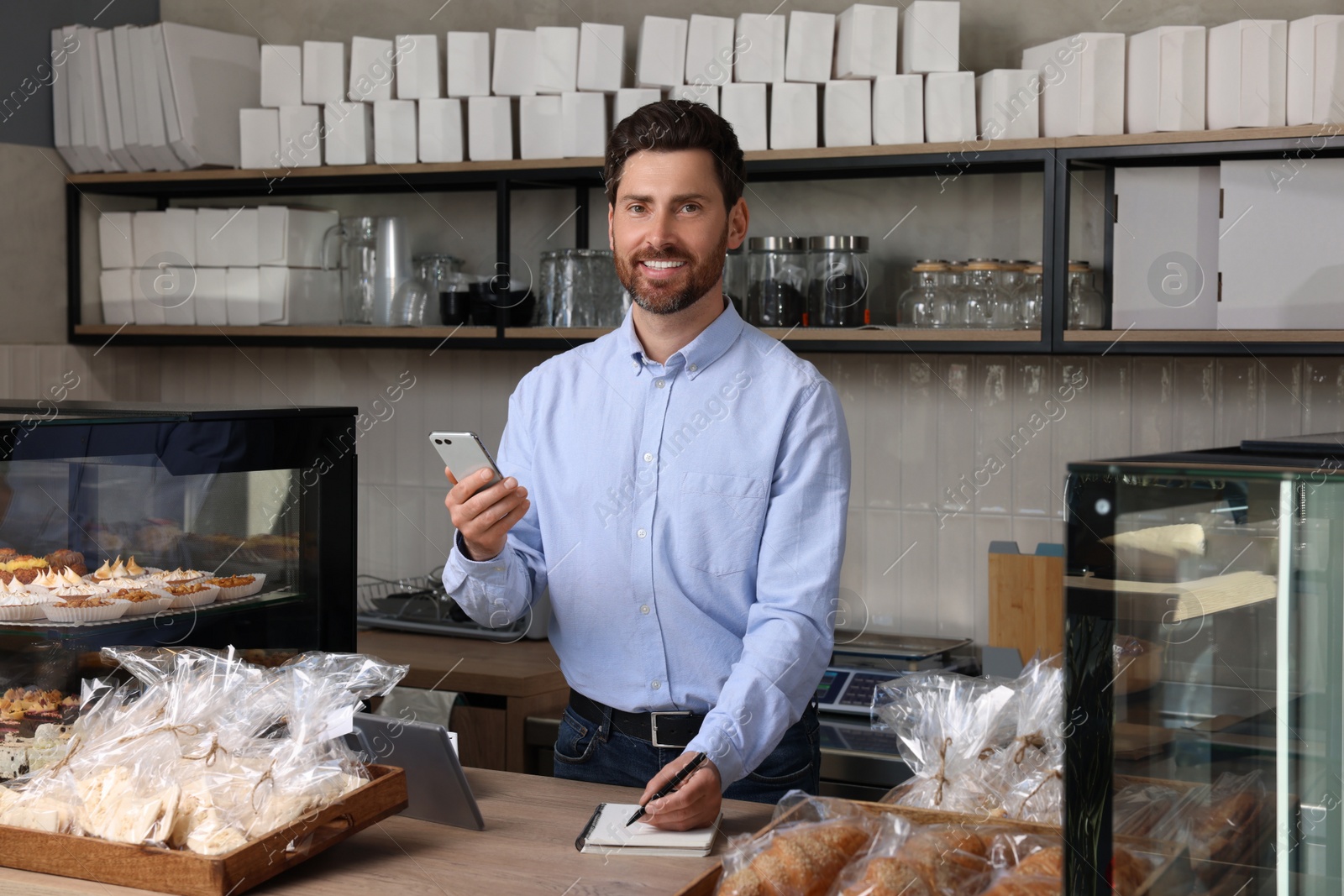 Photo of Happy business owner with smartphone and pen at cashier desk in bakery shop