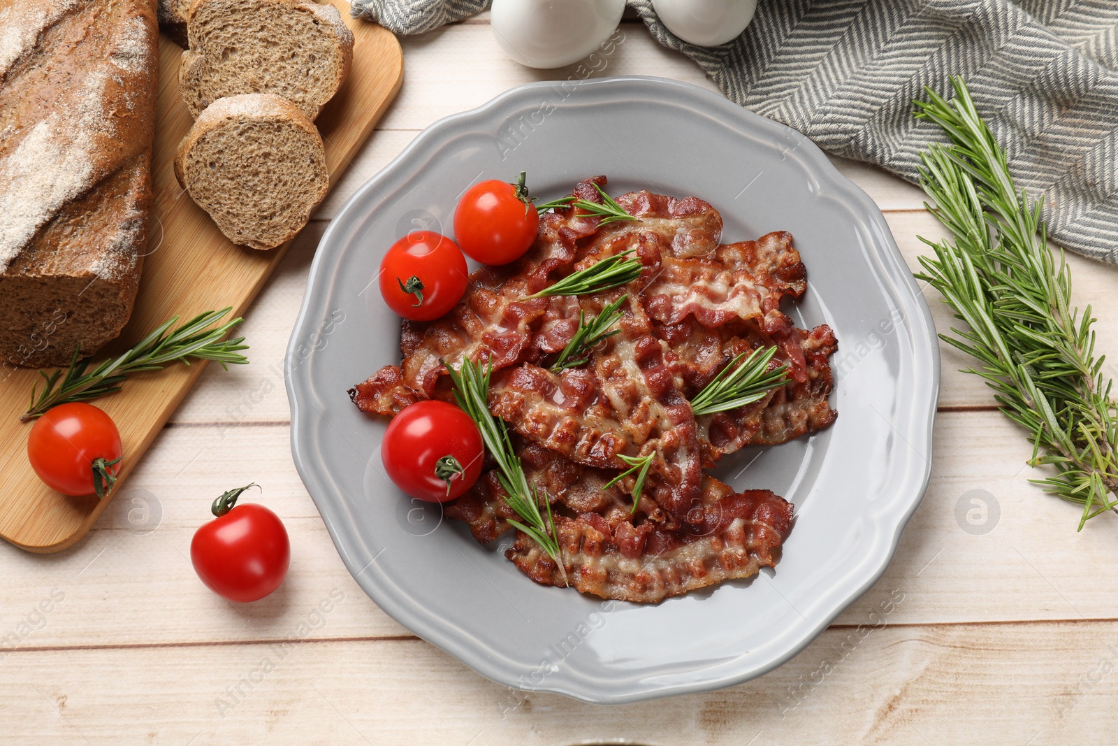 Photo of Slices of tasty fried bacon with rosemary and tomatoes served on wooden table, flat lay