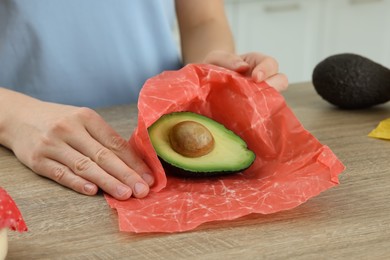 Photo of Woman packing half of avocado into beeswax food wrap at wooden table, closeup