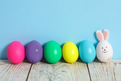 Photo of Easter eggs and white one as cute bunny on wooden table against light blue background