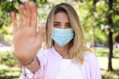 Photo of Woman in protective face mask showing stop gesture in park. Prevent spreading of coronavirus