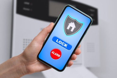 Woman operating home alarm system via mobile phone against white wall with security control panel, closeup. Application interface on device screen