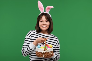 Photo of Easter celebration. Happy woman with bunny ears and wicker basket full of painted eggs on green background