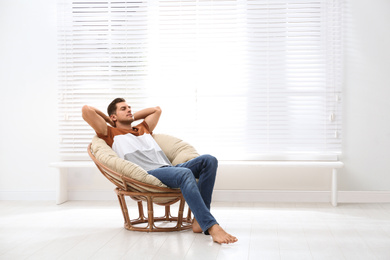 Attractive man relaxing in papasan chair near window at home. Space for text