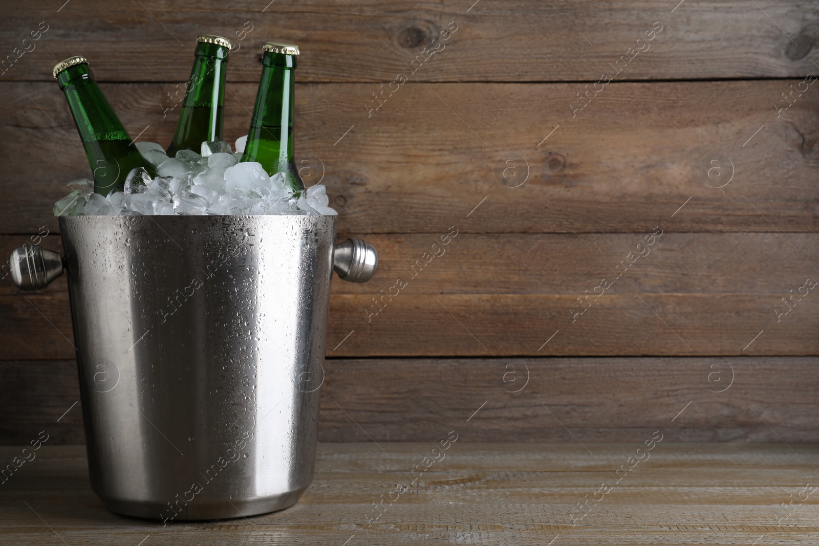 Photo of Metal bucket with beer and ice cubes on wooden table. Space for text