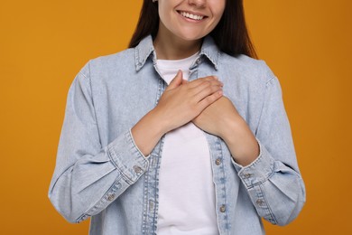 Photo of Thank you gesture. Grateful woman holding hands near heart on orange background, closeup