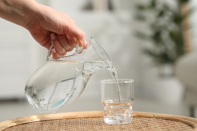 Photo of Woman pouring fresh water from jug into glass at wicker surface against blurred background, closeup