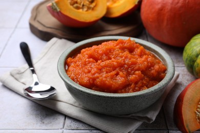 Photo of Bowl of delicious pumpkin jam and fresh pumpkin on tiled surface, closeup