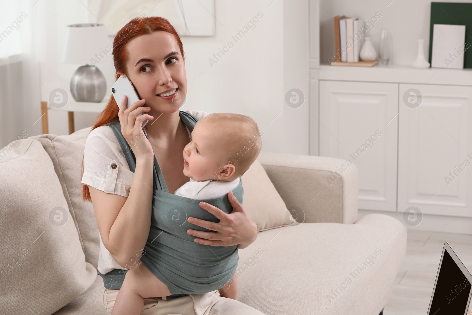 Photo of Mother talking on smartphone while holding her child in sling (baby carrier) at home