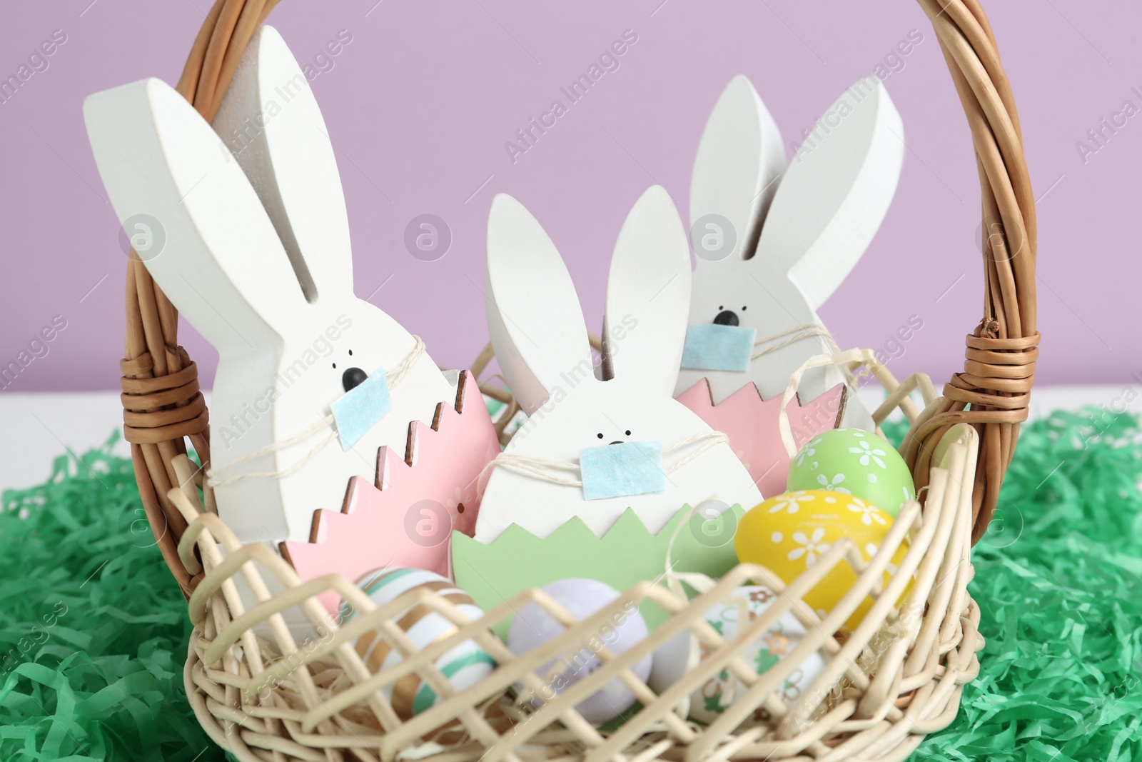 Photo of Wooden bunnies with protective masks and painted eggs in basket on paper grass, closeup. Easter holiday during COVID-19 quarantine
