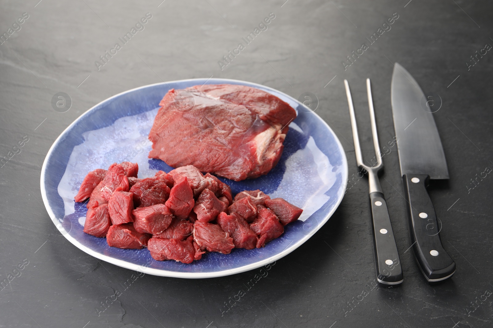 Photo of Plate with pieces of raw beef meat, knife and fork on grey textured table, closeup