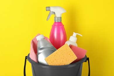 Photo of Bucket with different cleaning supplies against yellow background, closeup