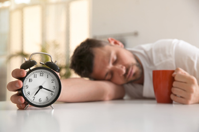 Photo of Man with alarm clock and cup of drink sleeping at table in morning, focus on hand