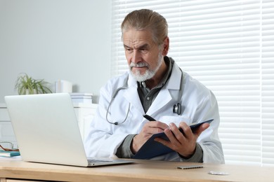 Senior doctor with laptop and clipboard consulting patient at wooden desk in clinic. Online medicine