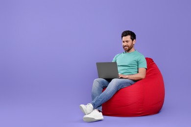 Happy man with laptop sitting on beanbag chair against purple background. Space for text