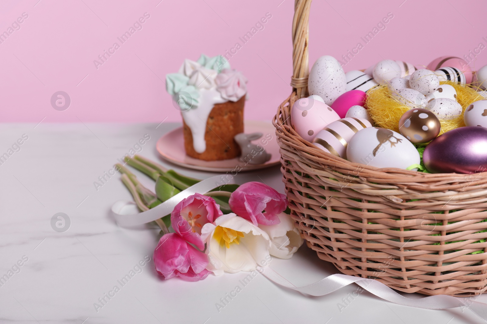 Photo of Wicker basket with festively decorated Easter eggs and beautiful tulips on white marble table against pink background. Space for text