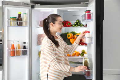 Photo of Young woman taking yoghurt out of refrigerator indoors