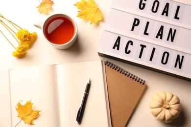 Photo of Light box with words Goal, Plan, Action, cup of tea, stationery and autumn decor on white table, flat lay