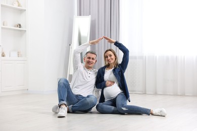 Photo of Young family housing concept. Pregnant woman with her husband forming roof with their hands while sitting on floor at home