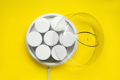 Modern yogurt maker with jars on yellow background, top view