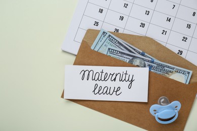 Photo of Maternity leave concept. Envelope with dollar banknotes, card, pacifier and calendar on beige background, flat lay. Space for text