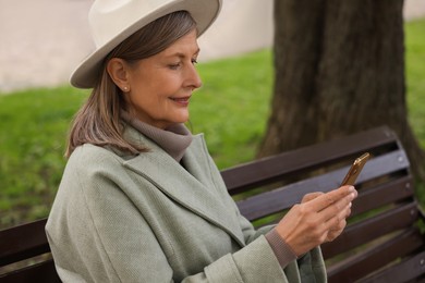 Beautiful senior woman sitting on bench and using smartphone outdoors, space for text