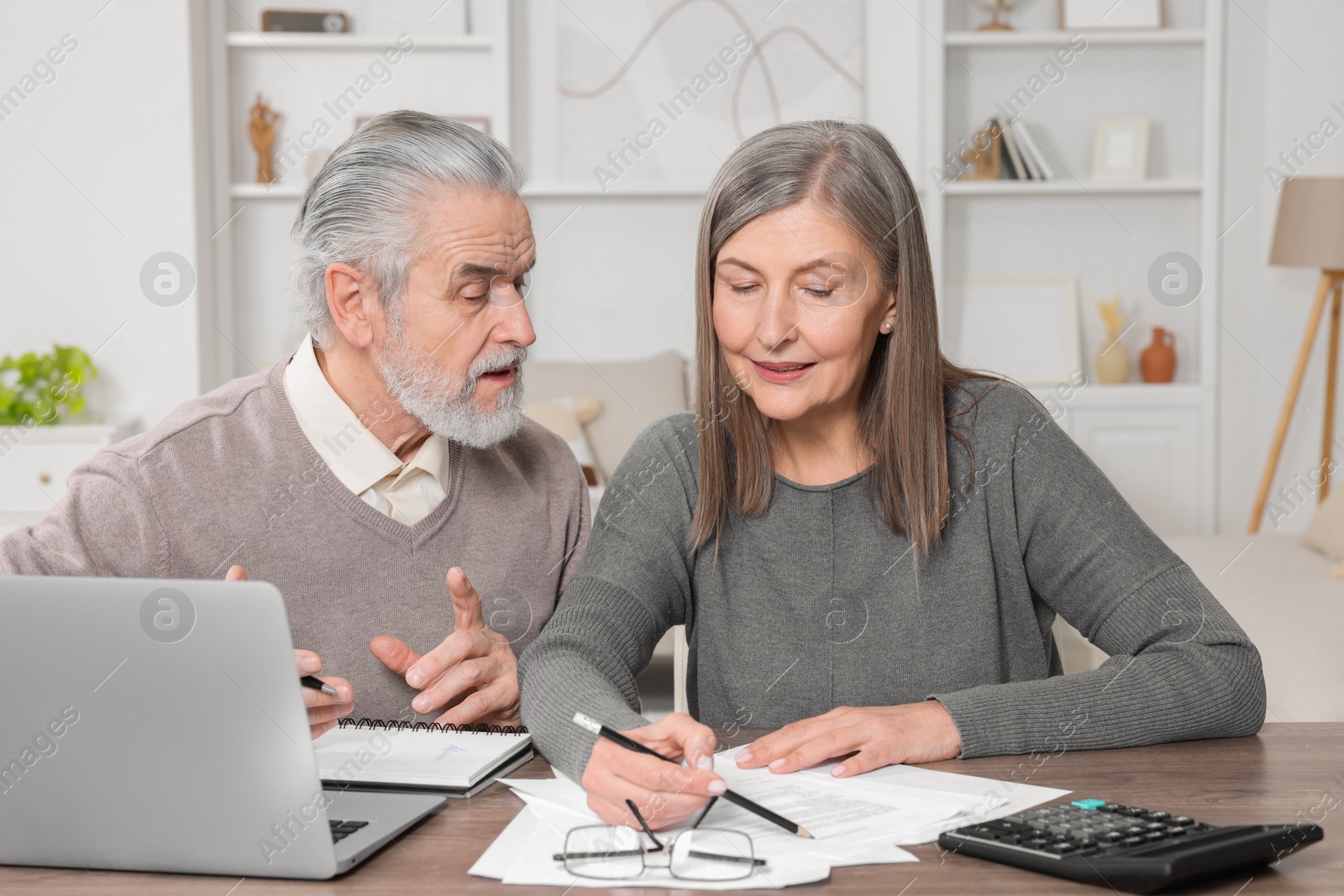 Photo of Elderly couple with papers and laptop discussing pension plan at wooden table in room