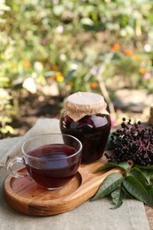Photo of Elderberry jam, glass cup of tea and Sambucus berries on table outdoors, space for text