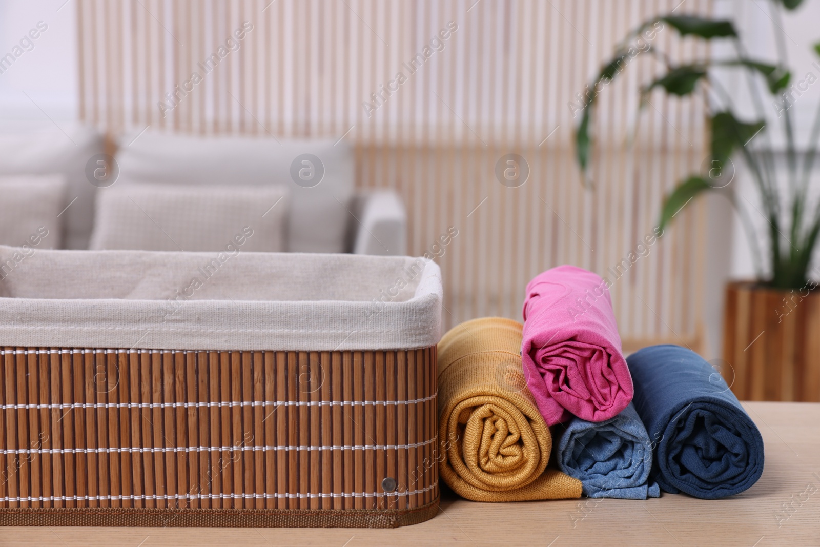 Photo of Rolled shirts near empty basket on table indoors. Organizing clothes