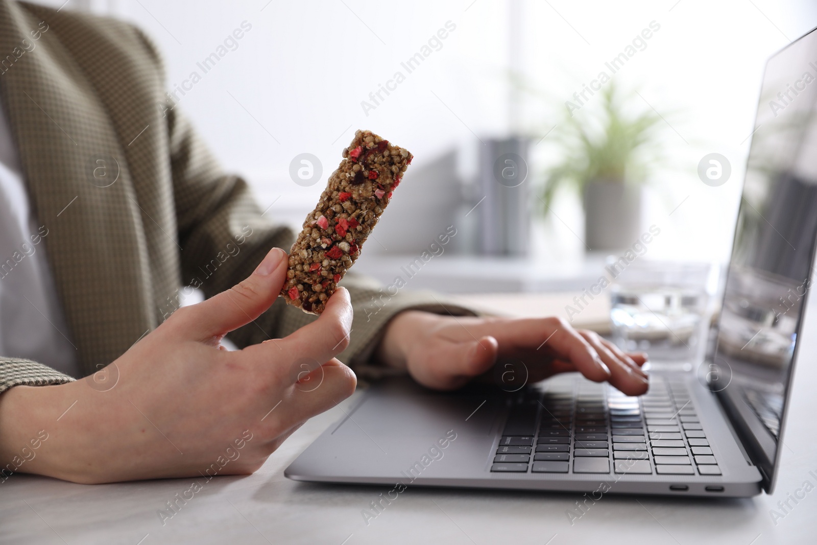Photo of Woman holding tasty granola bar working with laptop at light table in office, closeup