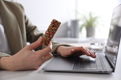 Photo of Woman holding tasty granola bar working with laptop at light table in office, closeup