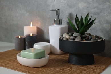 Potted artificial plant, soap, burning candles and bamboo mat on white table