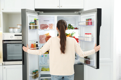 Photo of Young woman opening refrigerator in kitchen, back view