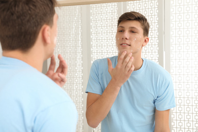 Teen guy with acne problem applying cream near mirror indoors