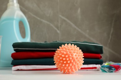 Photo of Orange dryer ball near stacked clean clothes, laundry detergents on white table