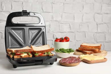 Photo of Modern grill maker with tasty sandwiches and ingredients on white wooden table