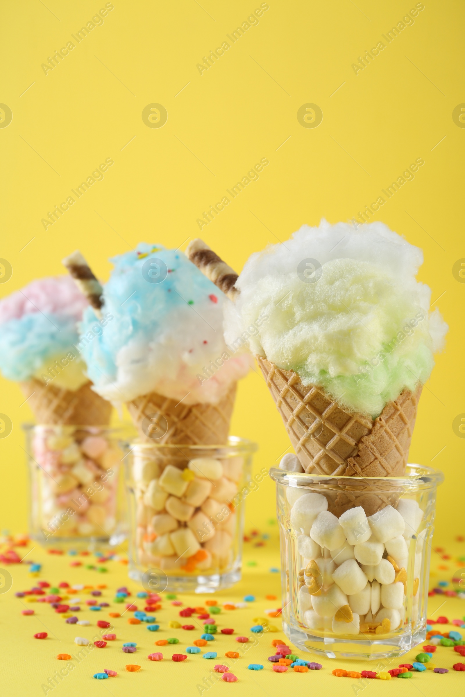 Photo of Sweet cotton candies in waffle cones and marshmallows on yellow background, closeup