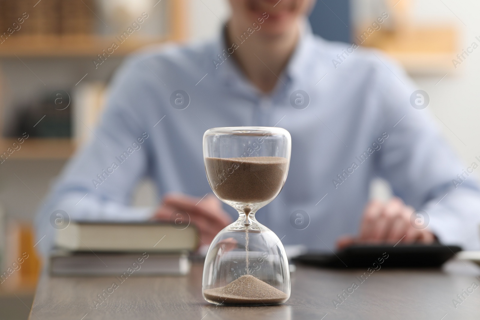 Photo of Hourglass with flowing sand on desk. Man using calculator indoors, selective focus