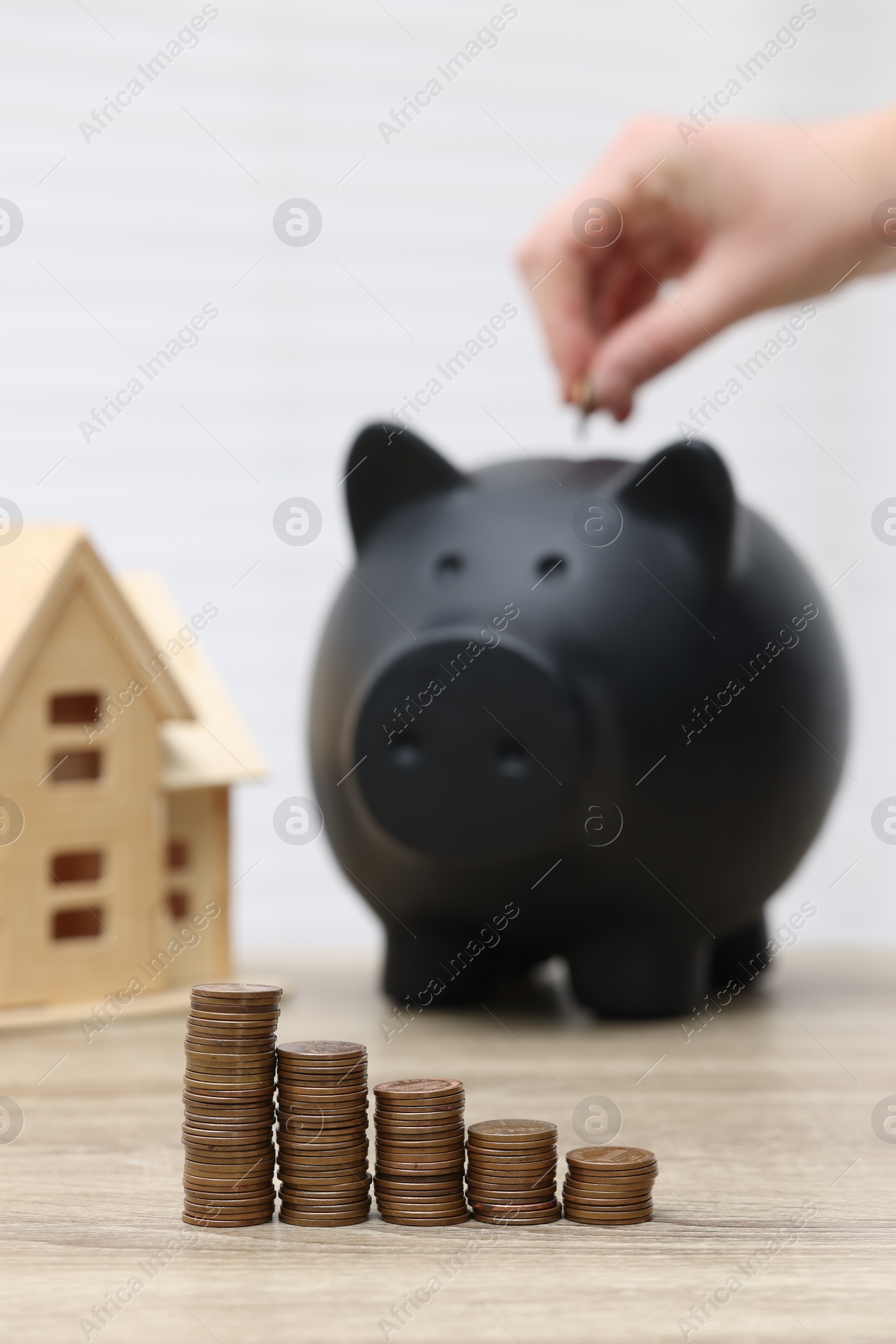 Photo of Woman putting coin into piggy bank at wooden table, focus on stacked coins