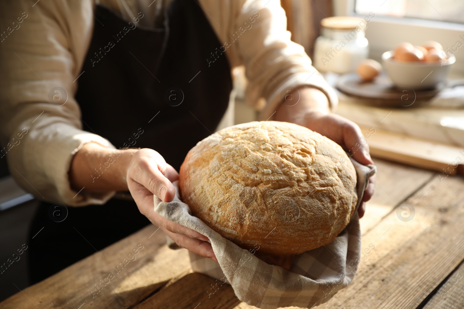 Photo of Man holding loaf of fresh bread at wooden table indoors, closeup